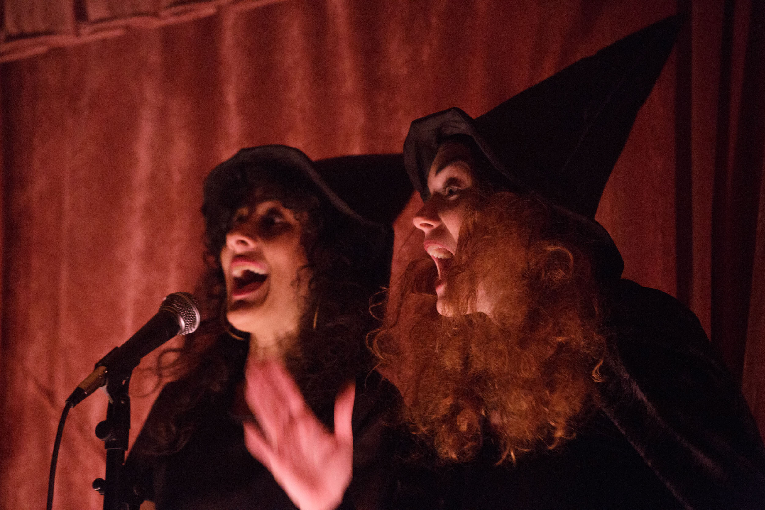 Two people dressed in black pointy hats animatedly speaking into a mic in front of a red curtain