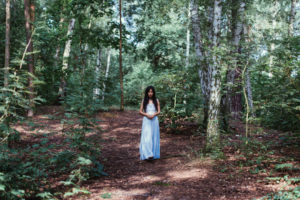 A person in a long blue dress with long black hair standing in a sun dappled forest