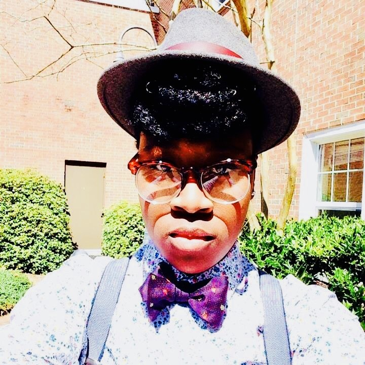 An image of Dani standing outside in the sun with a hat on and a blue shirt with a purple bowtie