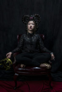 a figure in dark clothing against a black background, sitting is holding a crown