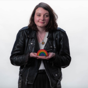 Grace Kredell standing against a white background wearing dark clothing holding a small wooden rainbow